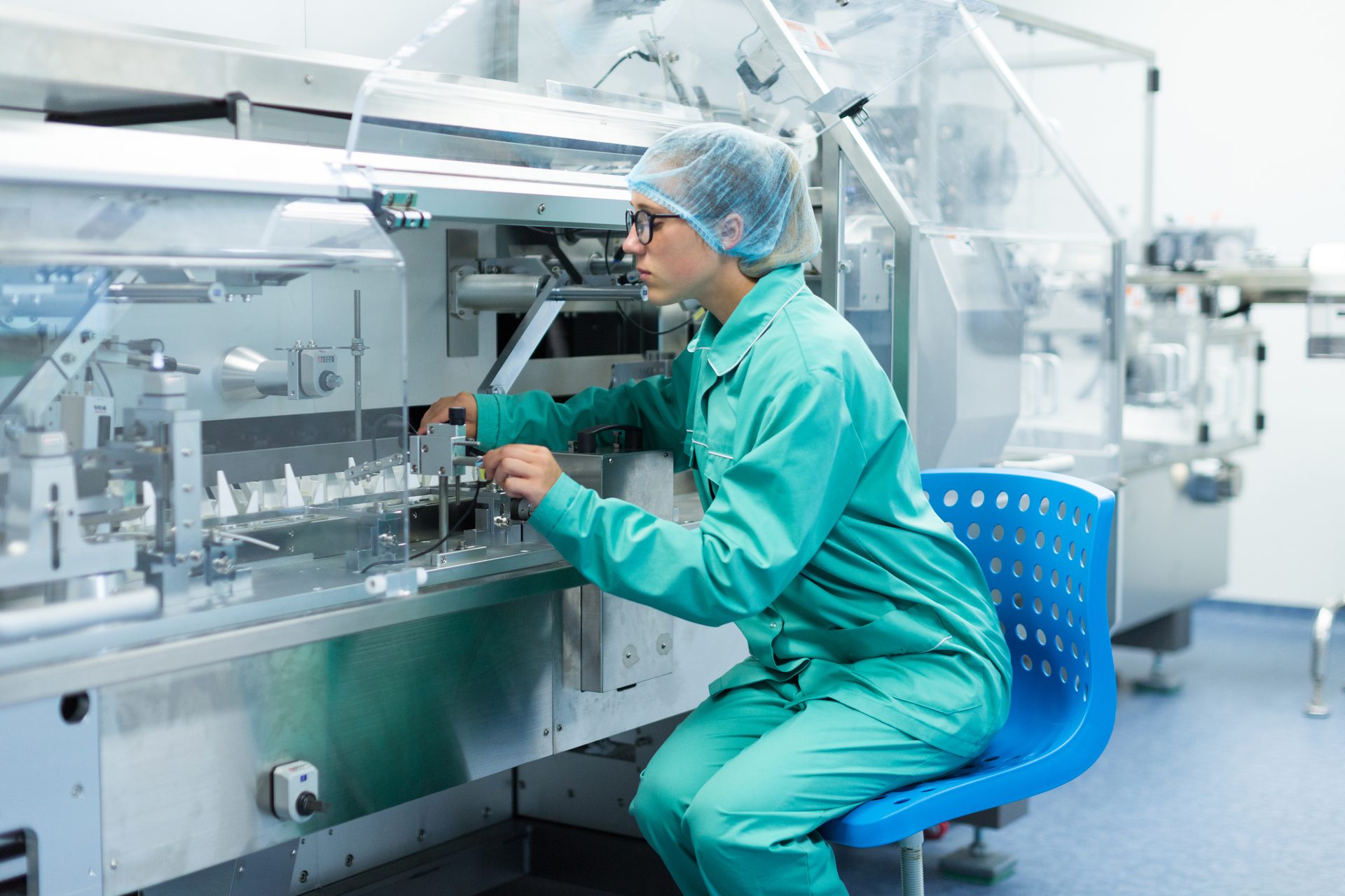 Technician in green coveralls and hairnet operating a stainless-steel packaging machine in a cleanroom