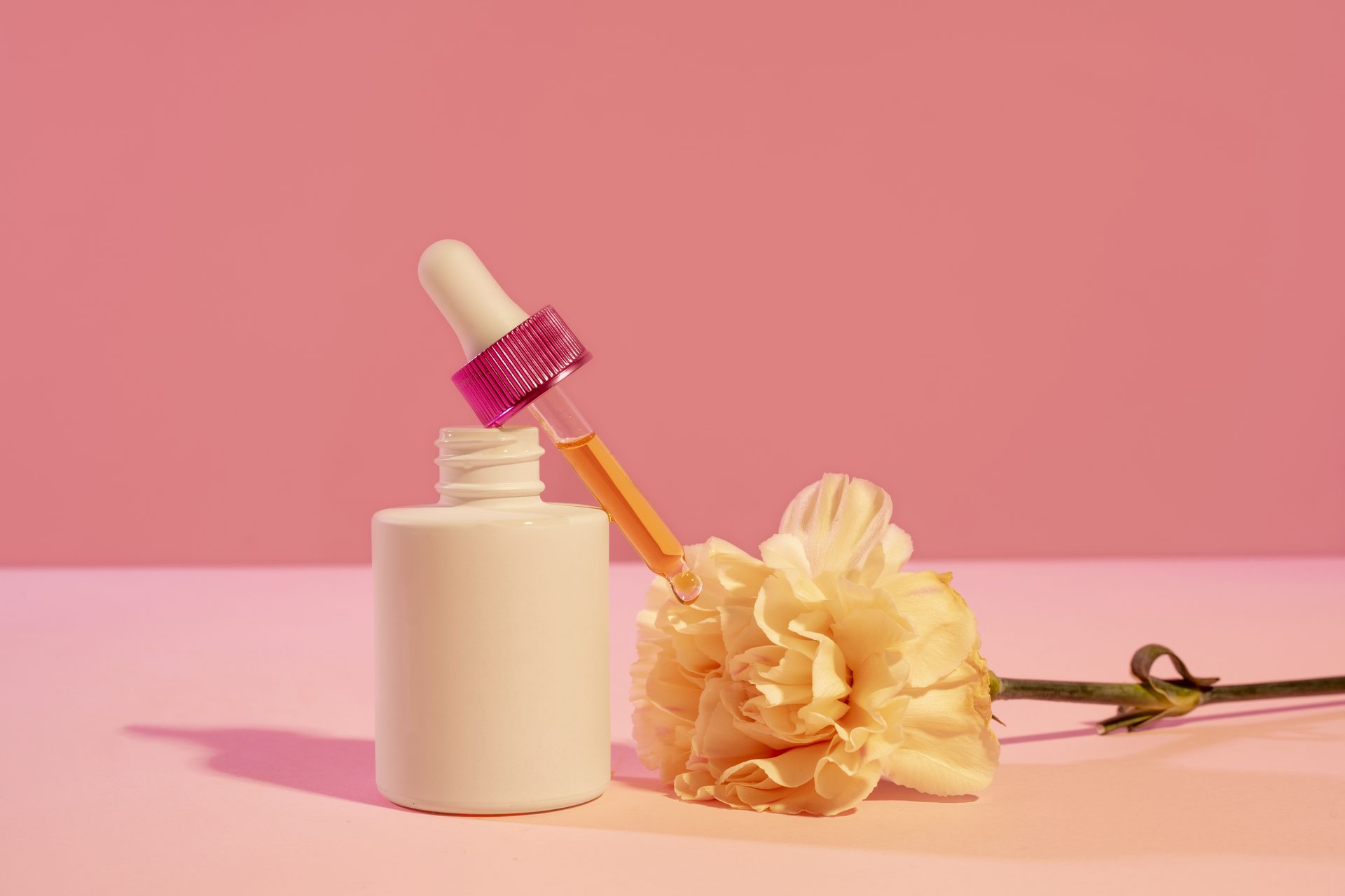 White dropper bottle with a pink cap beside a pale yellow carnation on a pink background