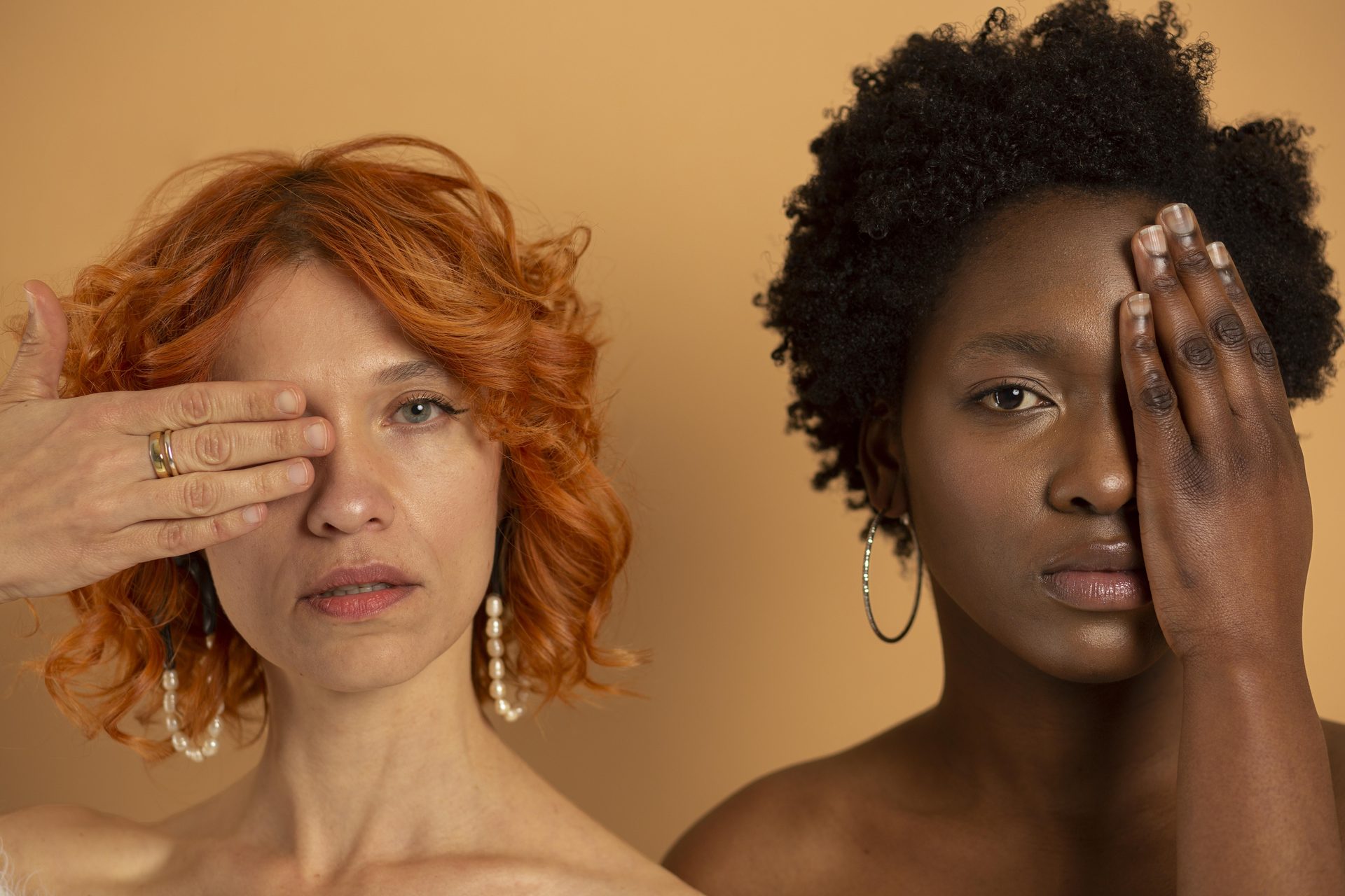 Two women on a tan backdrop each covering one eye by hand, one red-haired, one with afro hair