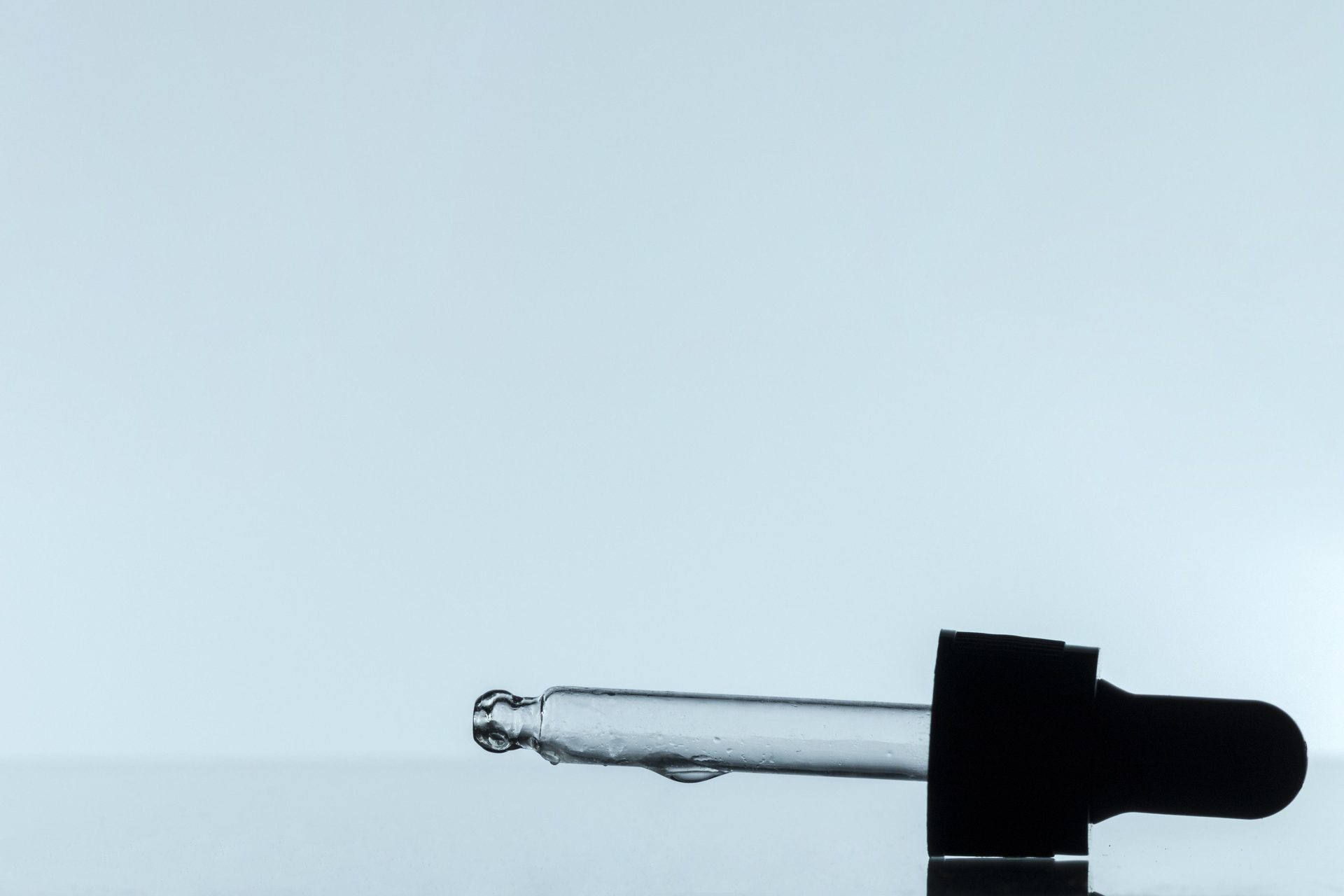 Glass dropper pipette with a black rubber bulb lying on its side against a pale blue-grey background