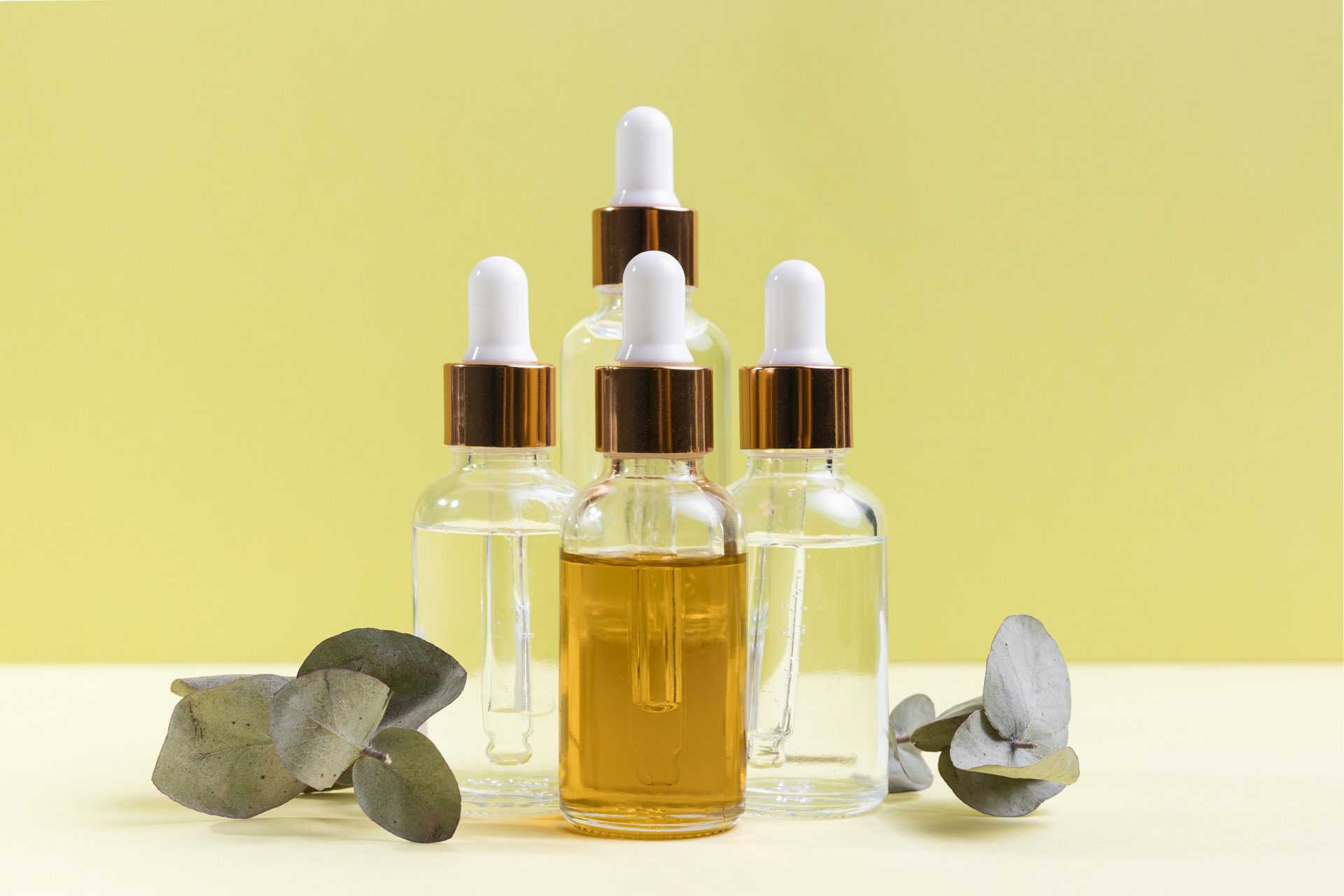 Five glass dropper bottles, one with amber oil, with eucalyptus leaves on a yellow background