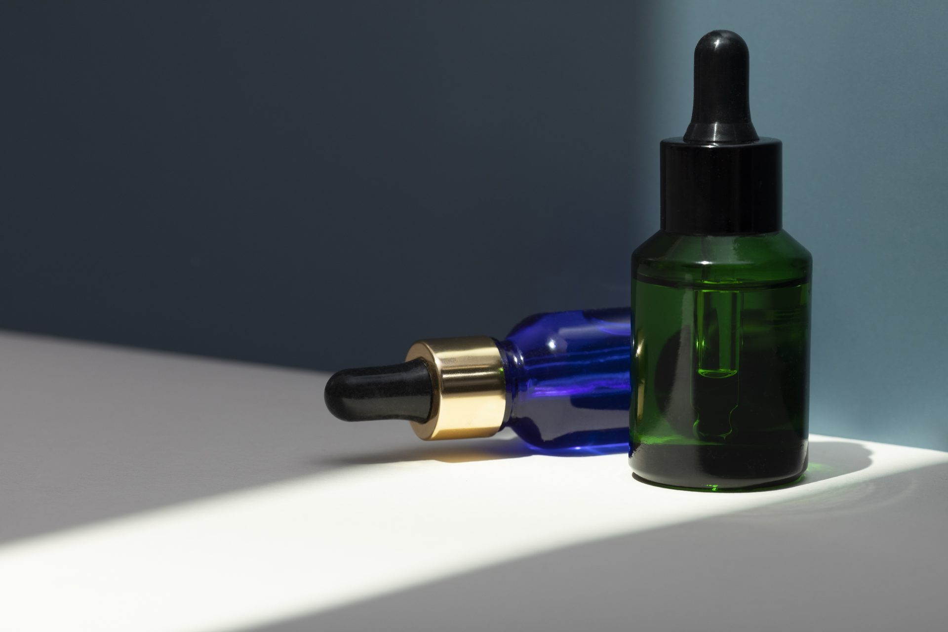Green glass dropper bottle upright and a blue one lying down in a beam of sunlight