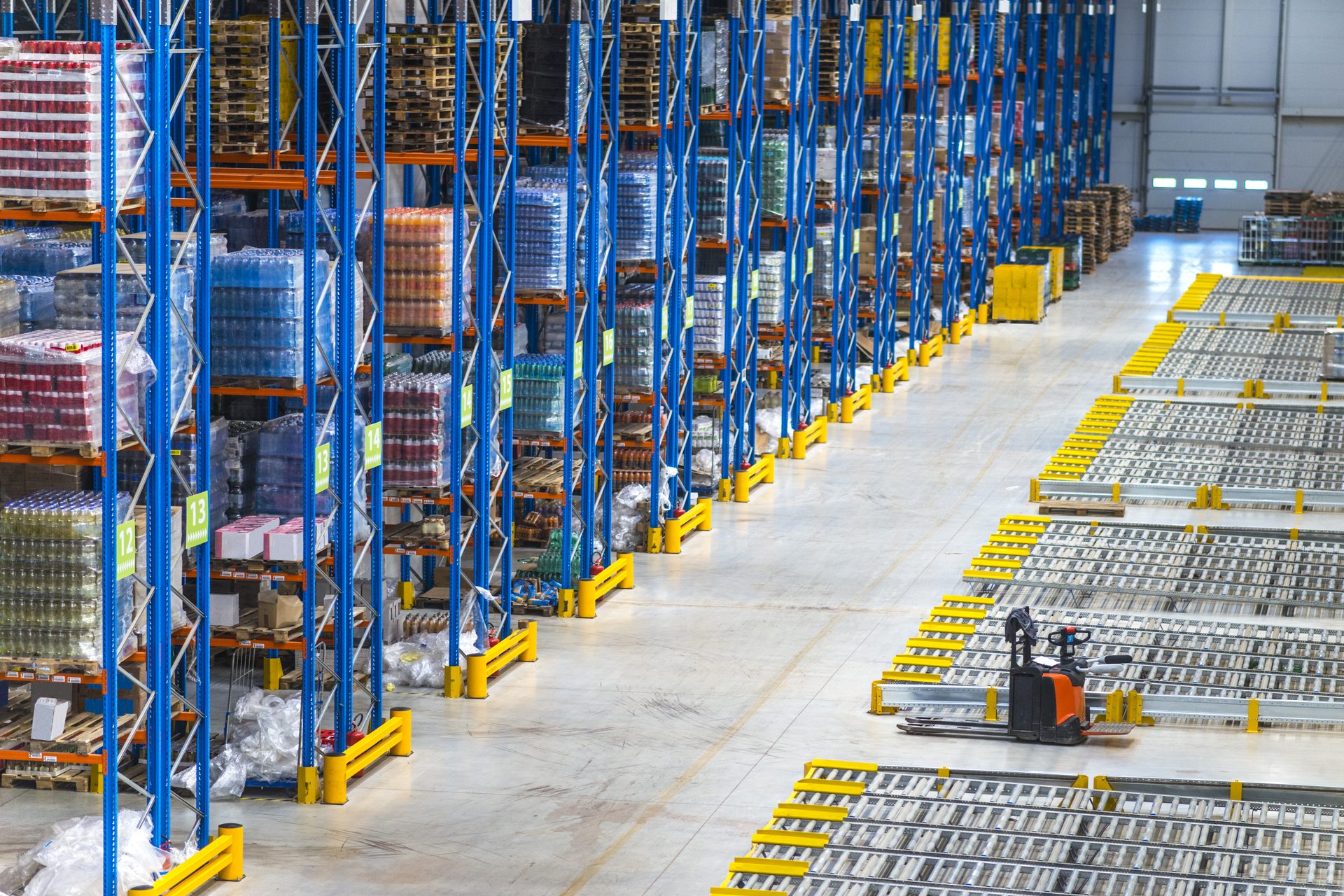Large warehouse with tall blue pallet racking, roller conveyors and an orange pallet truck