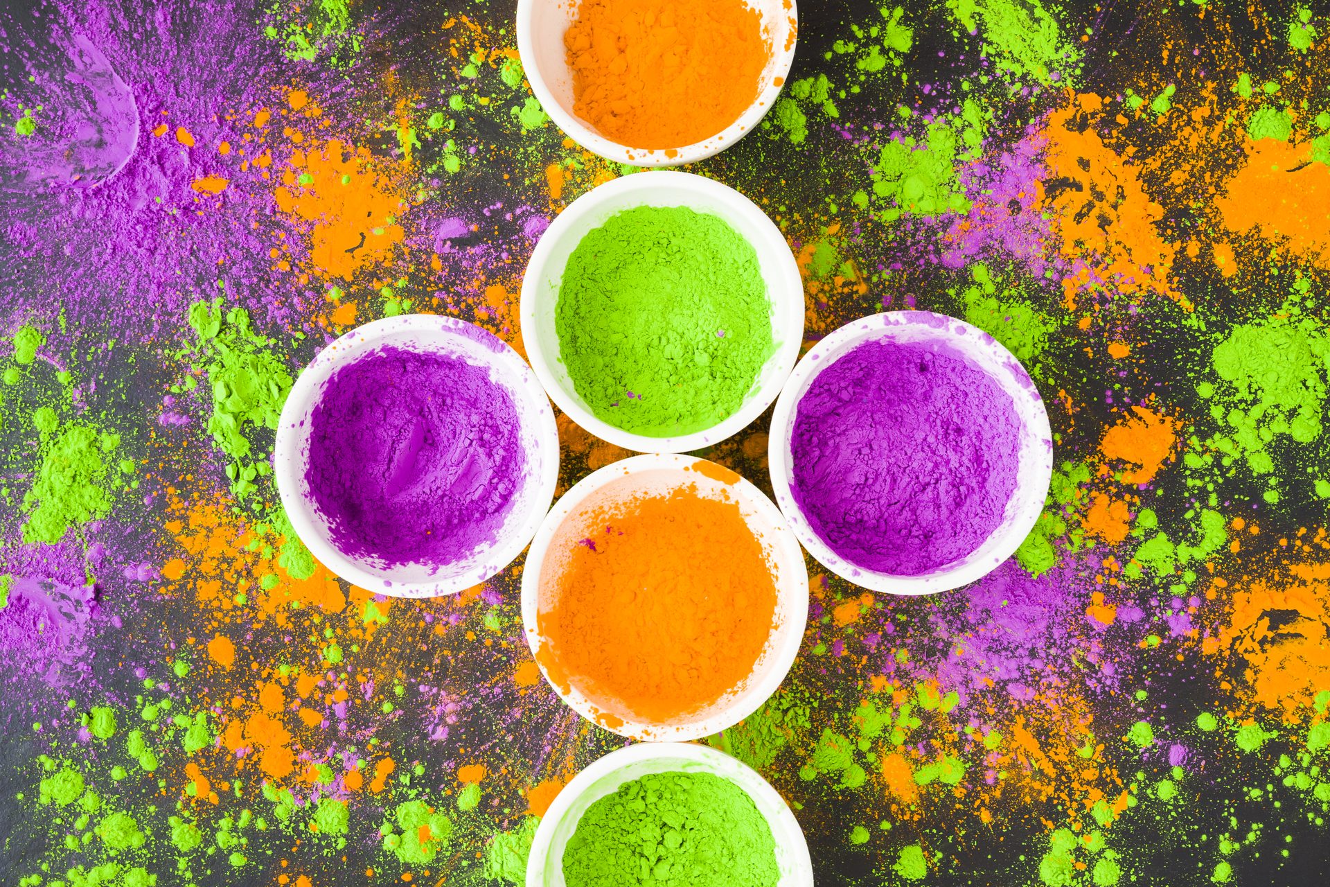 White bowls of orange, green and purple powder pigment amid scattered colour on a dark surface