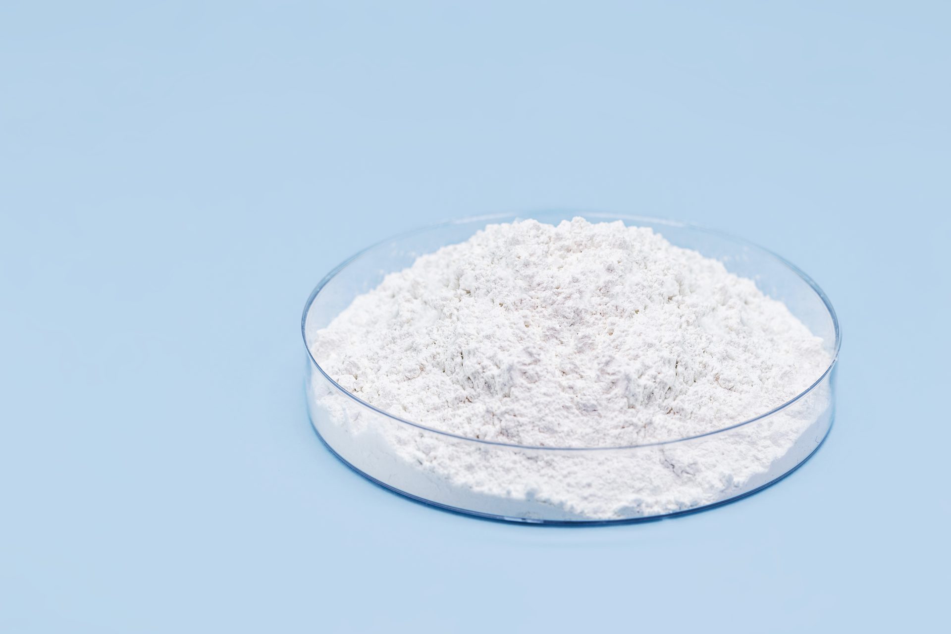 Glass petri dish heaped with fine white powder on a pale blue background