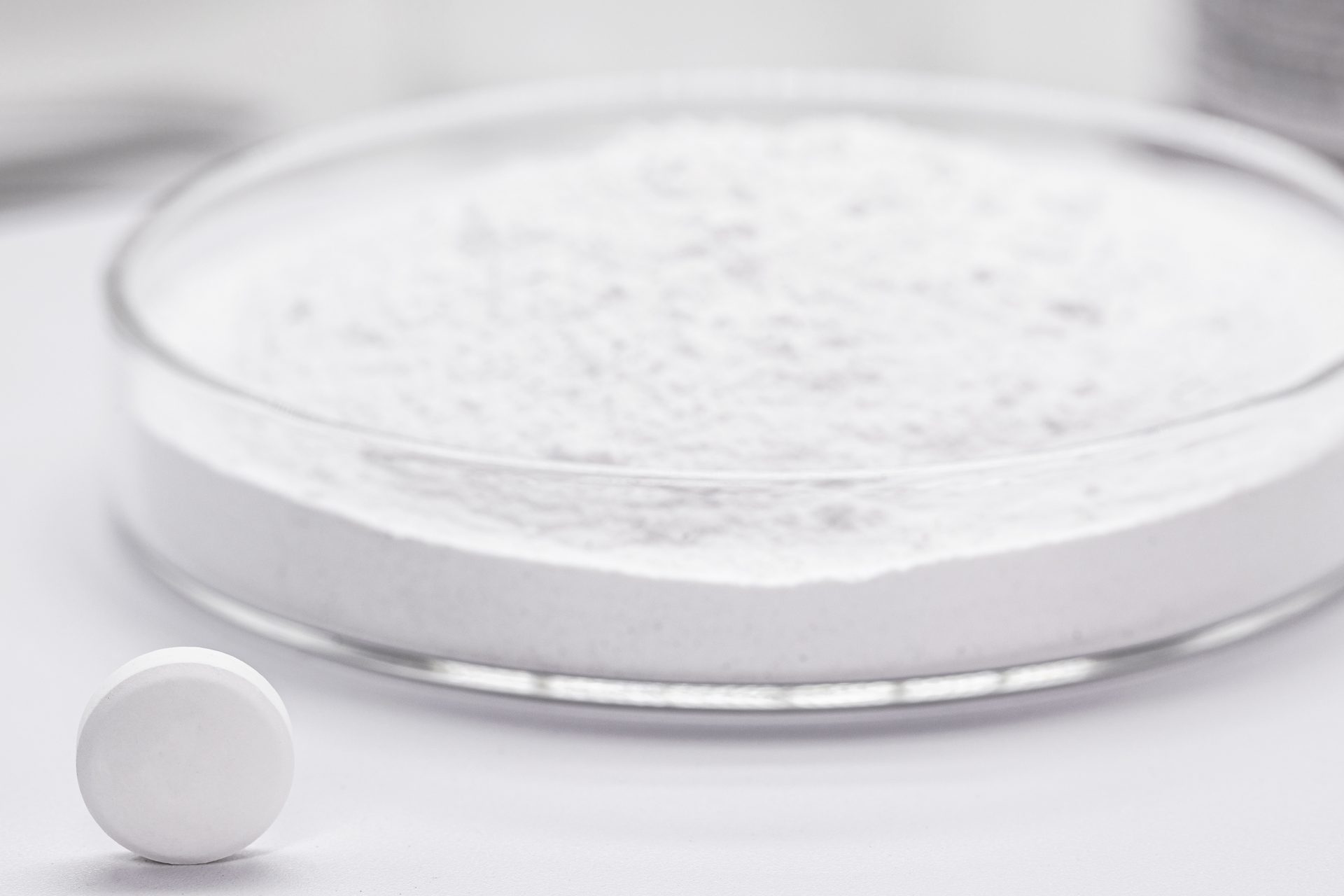 Glass dish filled with white powder beside a single white tablet on a white surface