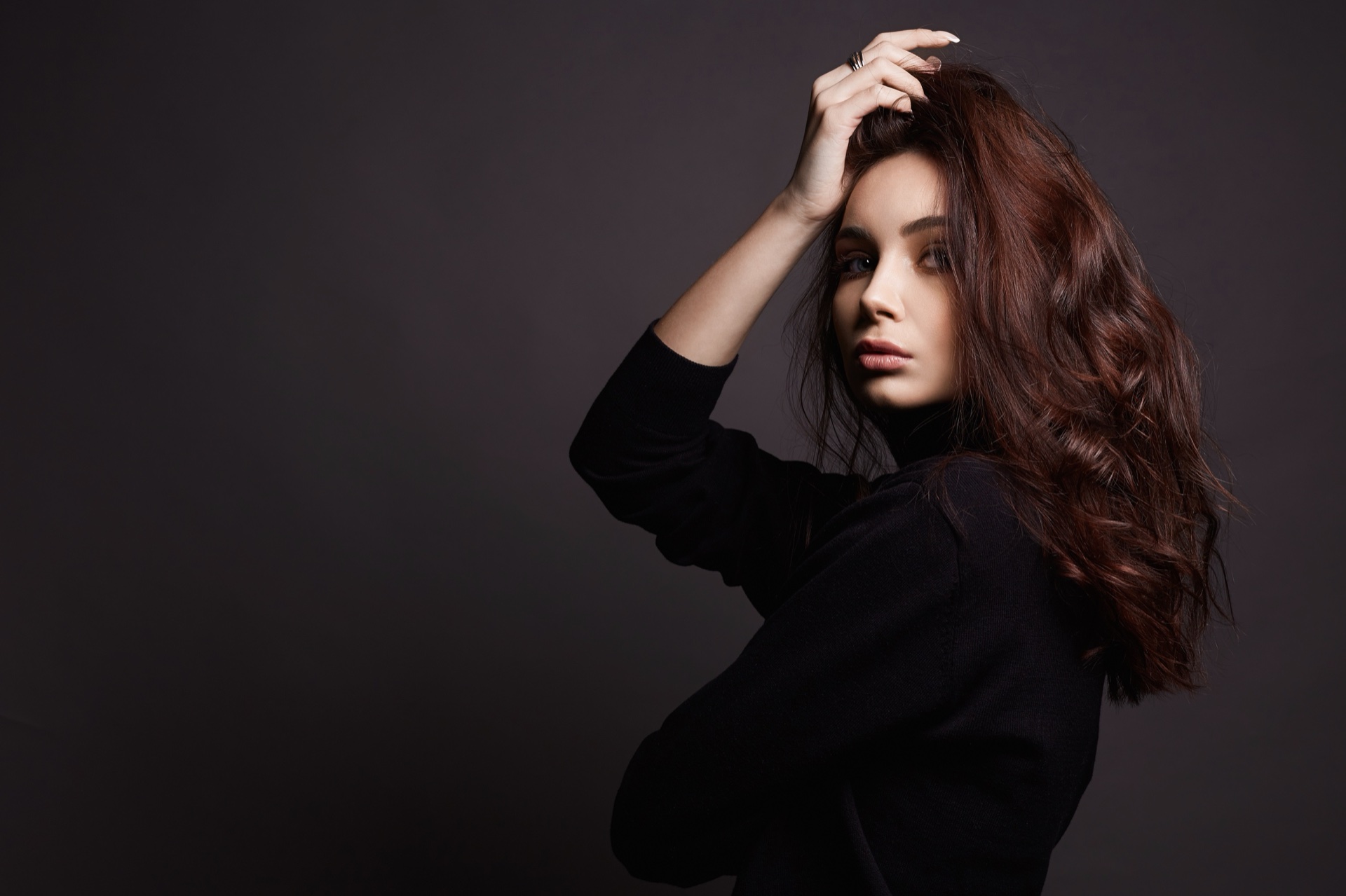 Woman with long wavy auburn hair in a black top, one hand raised to her head, dark backdrop