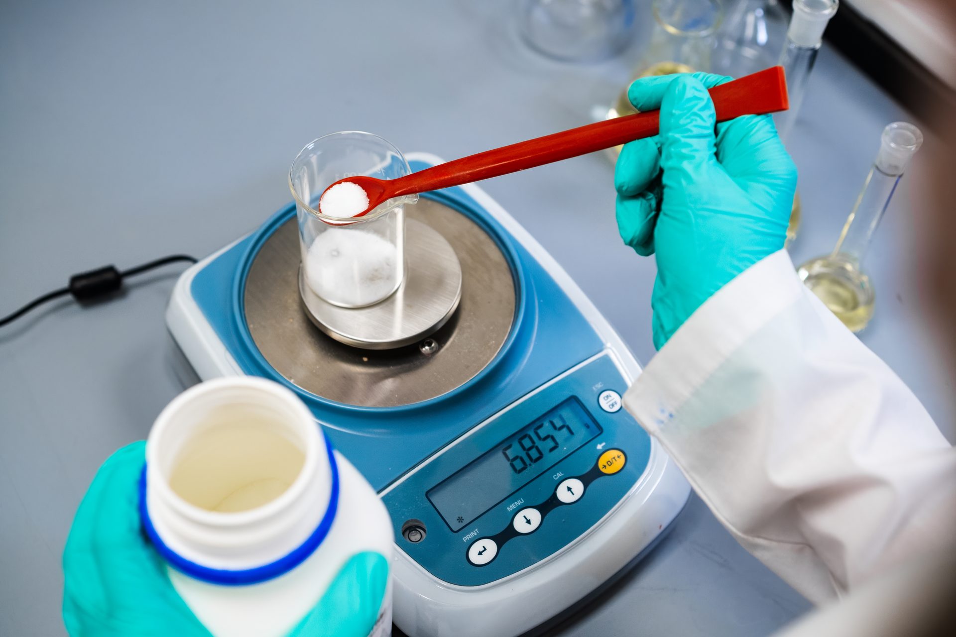 Gloved hands weighing white powder into a beaker on a digital balance using a red scoop