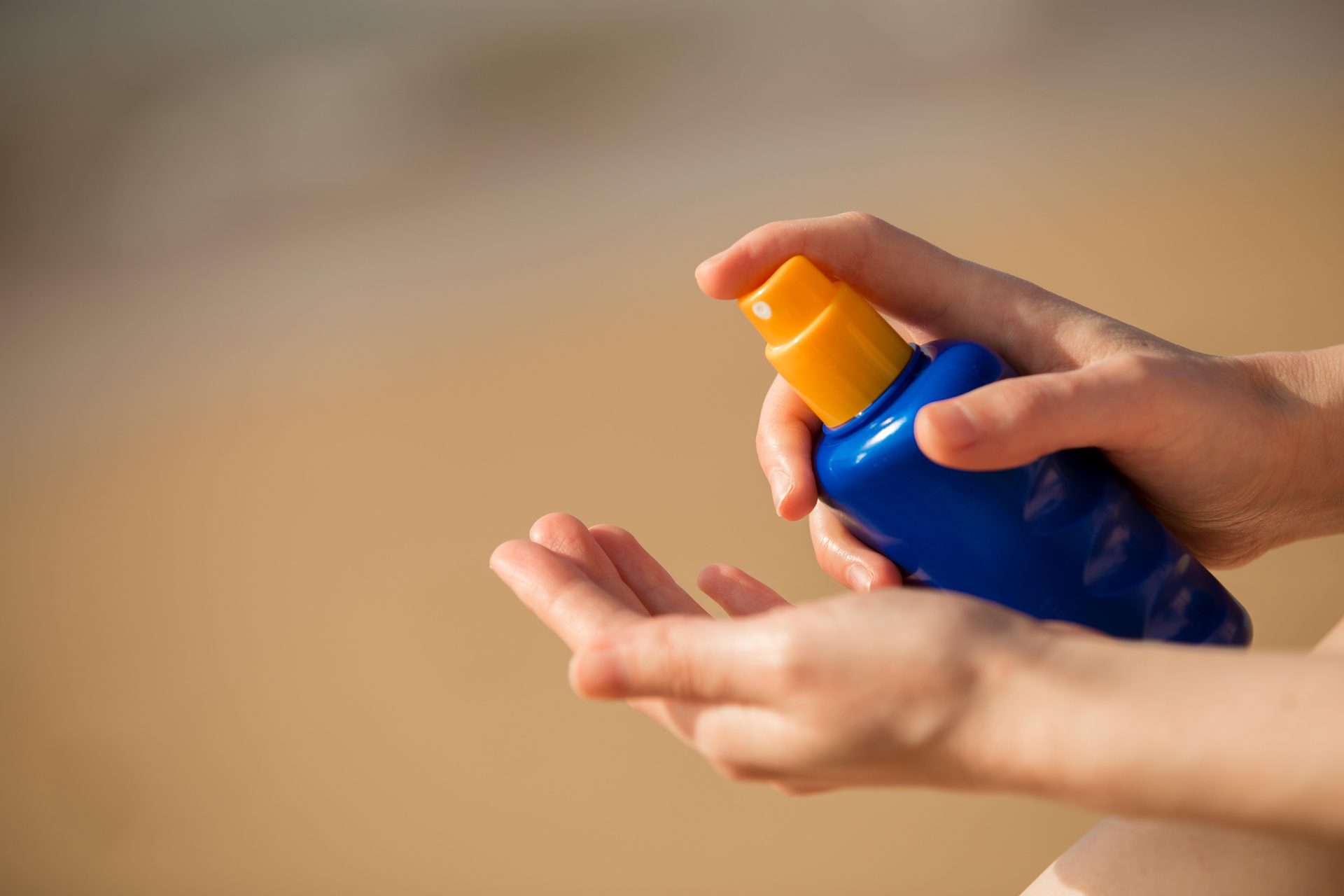 Hands holding a blue spray bottle with an orange cap over an open palm, sandy beach behind