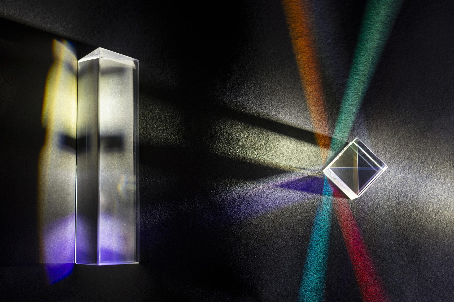 Two glass prisms on a dark surface casting coloured beams of refracted light
