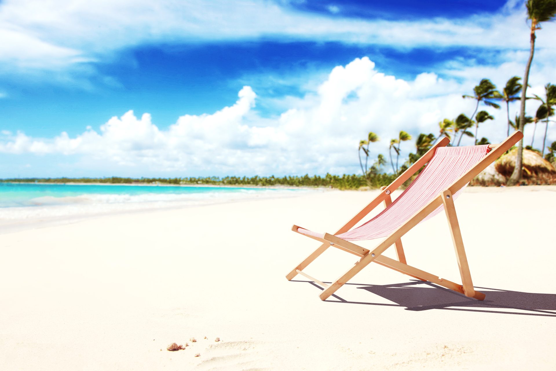 Wooden deckchair with pink striped fabric on white sand, palm trees and turquoise sea behind