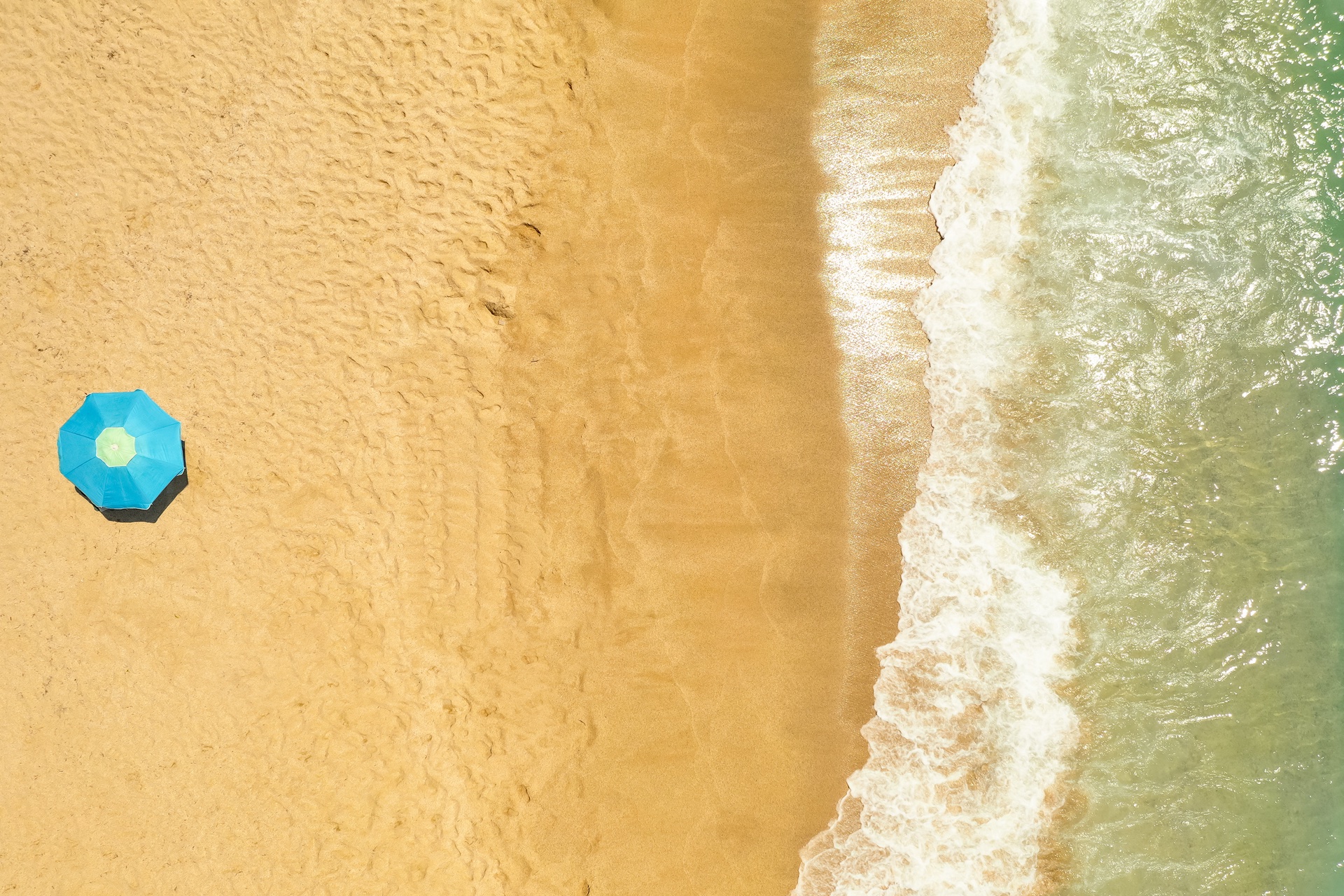 Aerial view of a golden sandy beach with a single blue parasol beside breaking turquoise waves