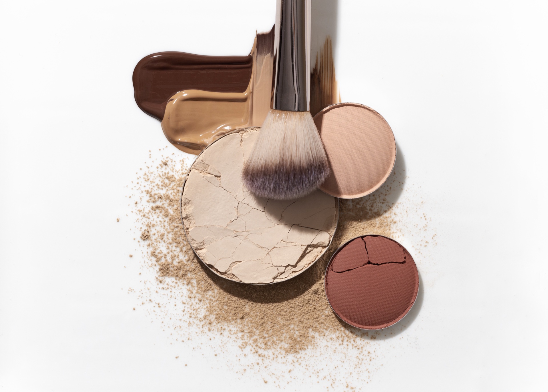 Cracked beige pressed powder, brush, loose powder and foundation smears on a white background